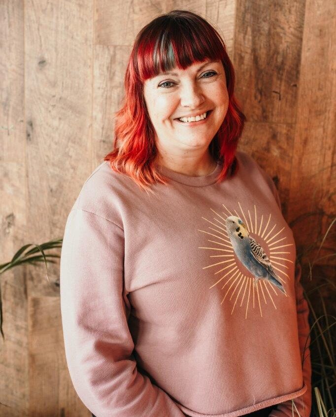 A white woman with read hair and a blue streak is smiling at the camera. She is wearing a pink sweatshirt with a budgie on it. She's against a wood background