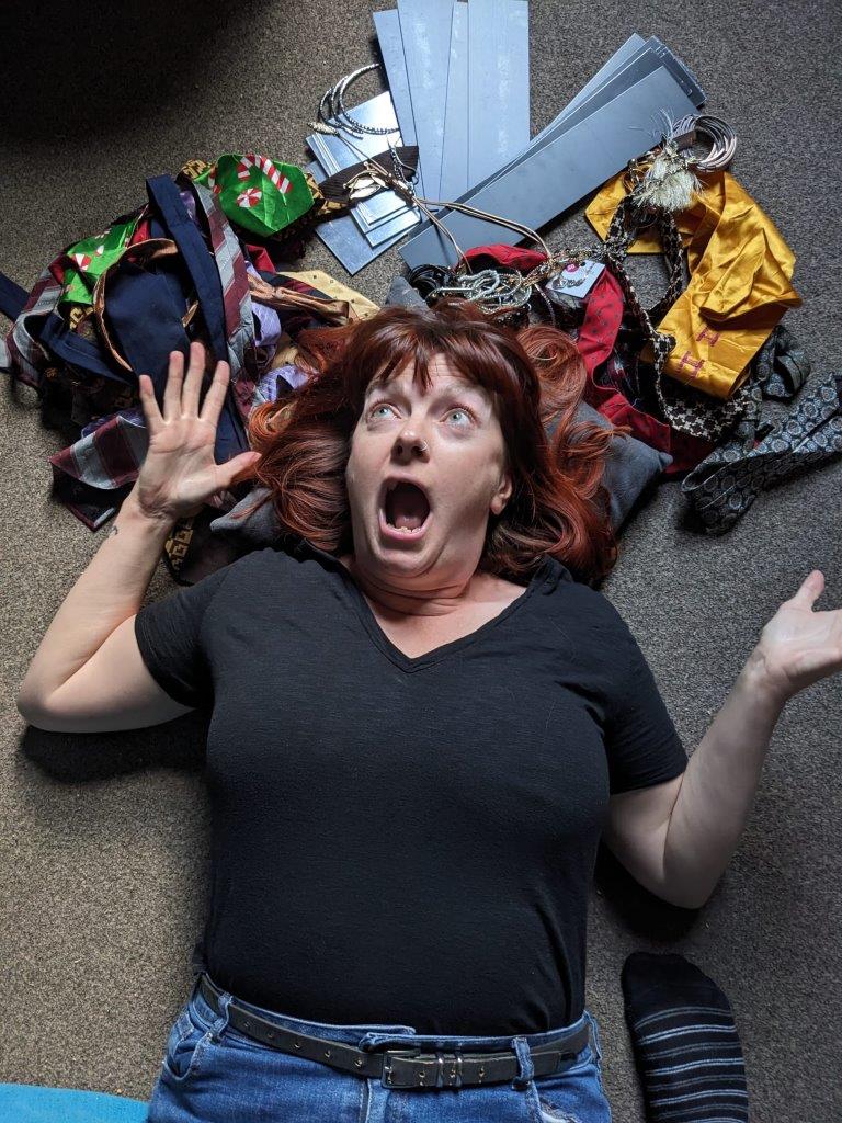 A white woman with dark hair is lying on the ground wearing a black tshirt and blue jeans. She is surrounded by waste textiles and has a shocked look on her face.