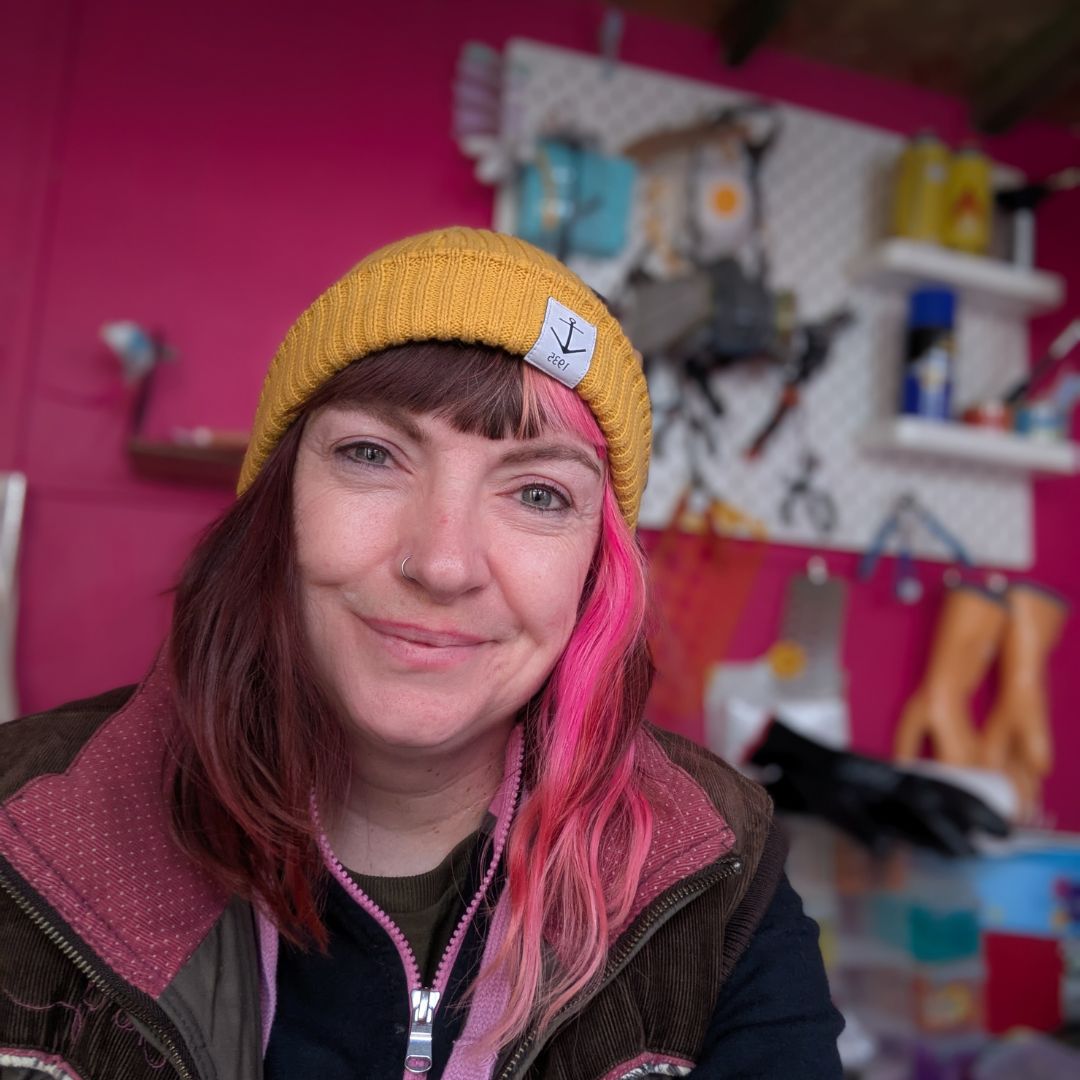 TP website Profile Photo – About Us A white woman with pink hair and a yellow hat poses for the camera in her workshop. The walls are pink and tools are hanging up.