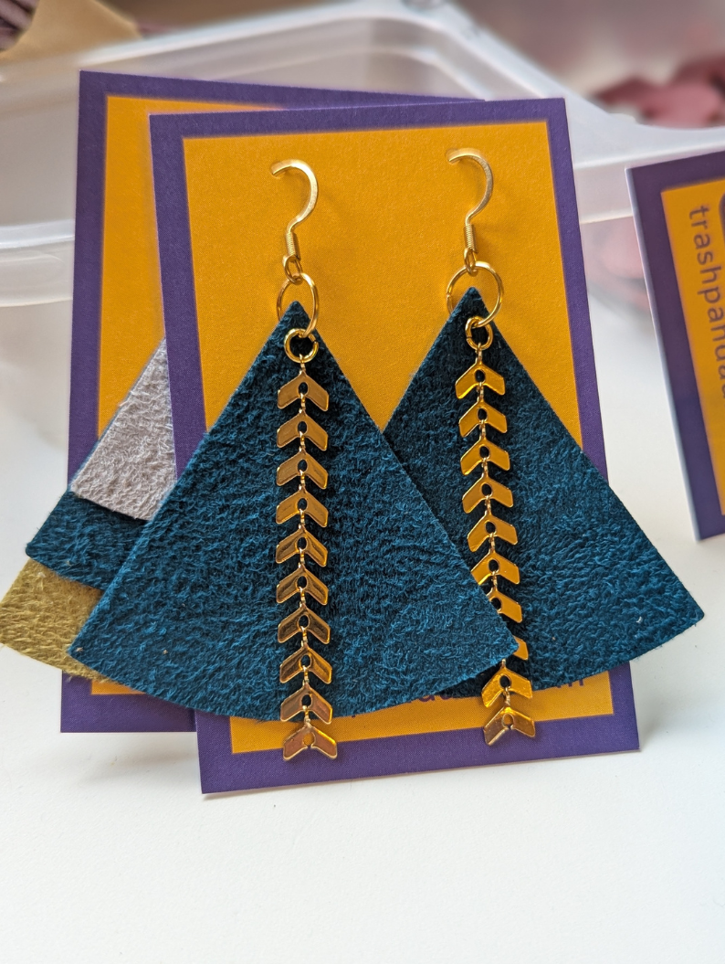 A pair of teal coloured triangle earrings sit on a jewellery display card. They are adorned with gold-coloured metal