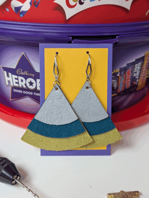 Tiered suedette triangle earrings in grey teal and mustard, set on a jewellery display card against some chocolate tubs