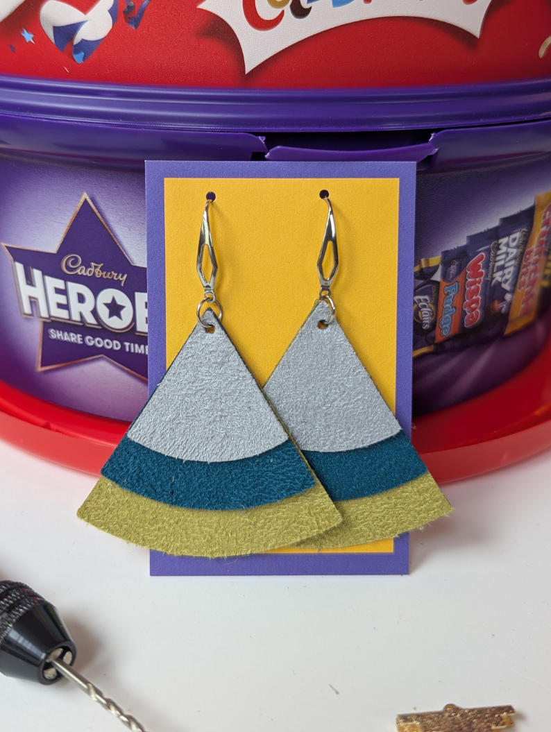 Tiered suedette triangle earrings in grey teal and mustard, set on a jewellery display card against some chocolate tubs