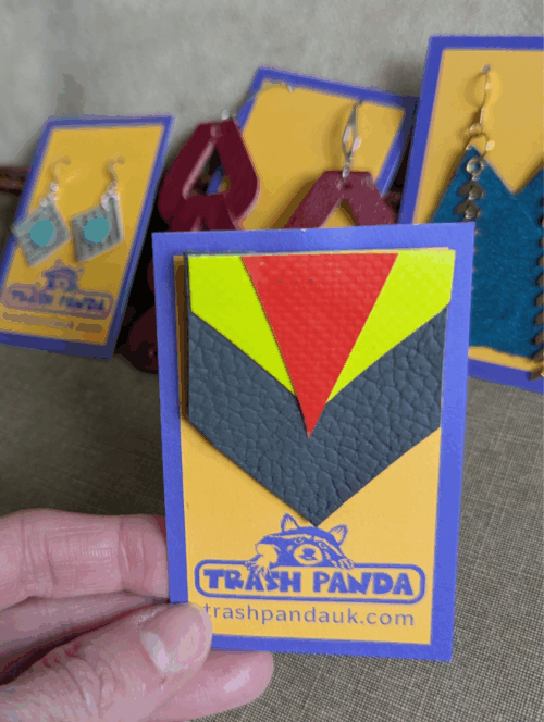 Geometric upcycled brooch with grey leather base and neon inflatable accents in red and yellow, displayed on Trash Panda backing card, held by a hand