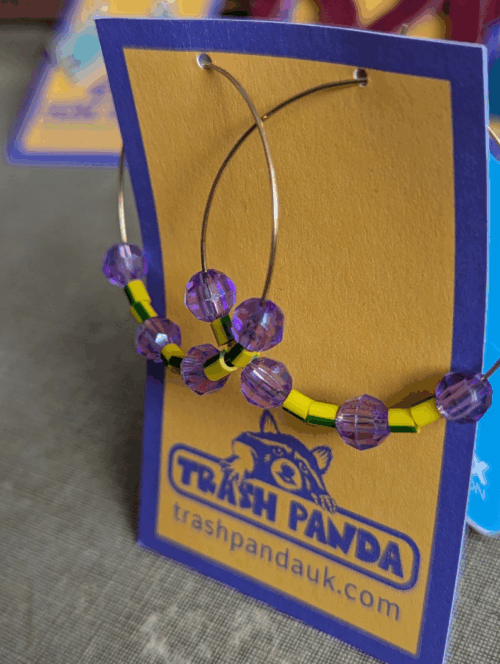Upcycled hoop earrings with yellow-and-green electrical wire beads and faceted purple beads on stainless steel hoops, displayed on Trash Panda card